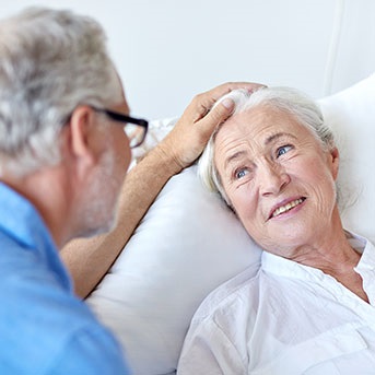 Seniorin liegt in Bett und Partner streichelt ihren Kopf