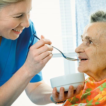 Pflegerin füttert Seniorin zur Aspirationsprophylaxe